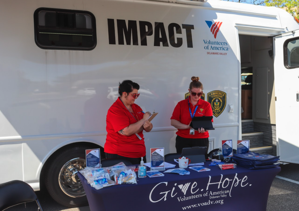 Two IMPACT staff members work on laptops and clipboards in front of a mobile IMPACT unit. 